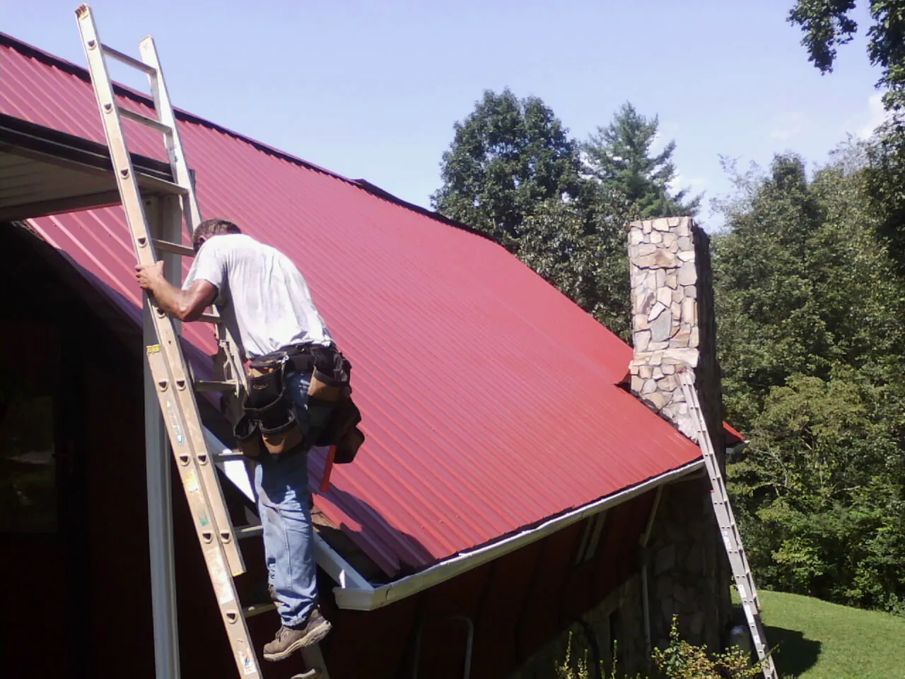 Roofer on a ladder beside a red metal roof and stone chimney — Soffit & Fascia Repair by Roofing Brothers in Bagdad
